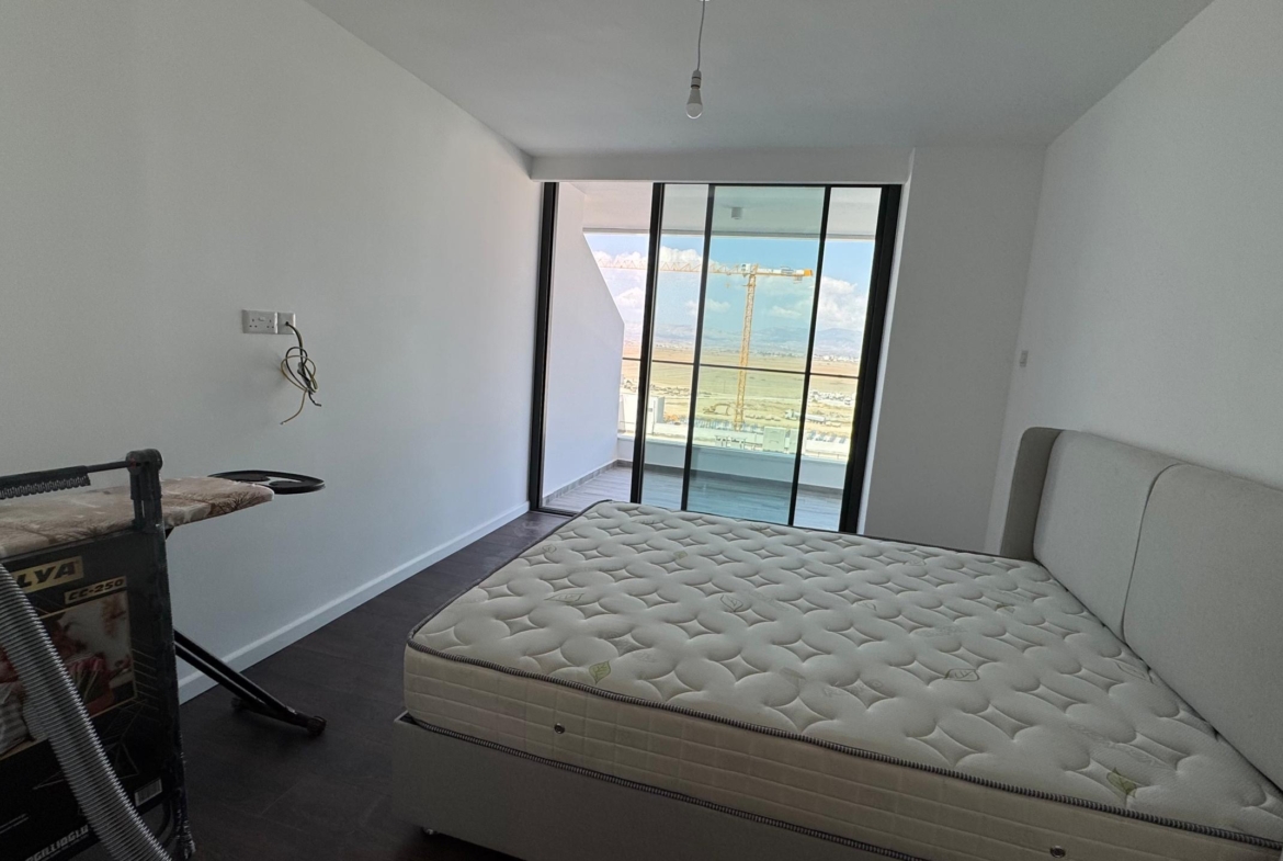 kıbrıs Grand Sapphire Resort & Residence Kıbrıs'ın minimal yatak odasında çıplak bir yatak, ütü masası, beyaz duvarlar, koyu zemin, çıplak ampul ve muhteşem dağ ve gökyüzü manzaralı bir balkona açılan büyük cam kapılar bulunmaktadır. aysan_giti