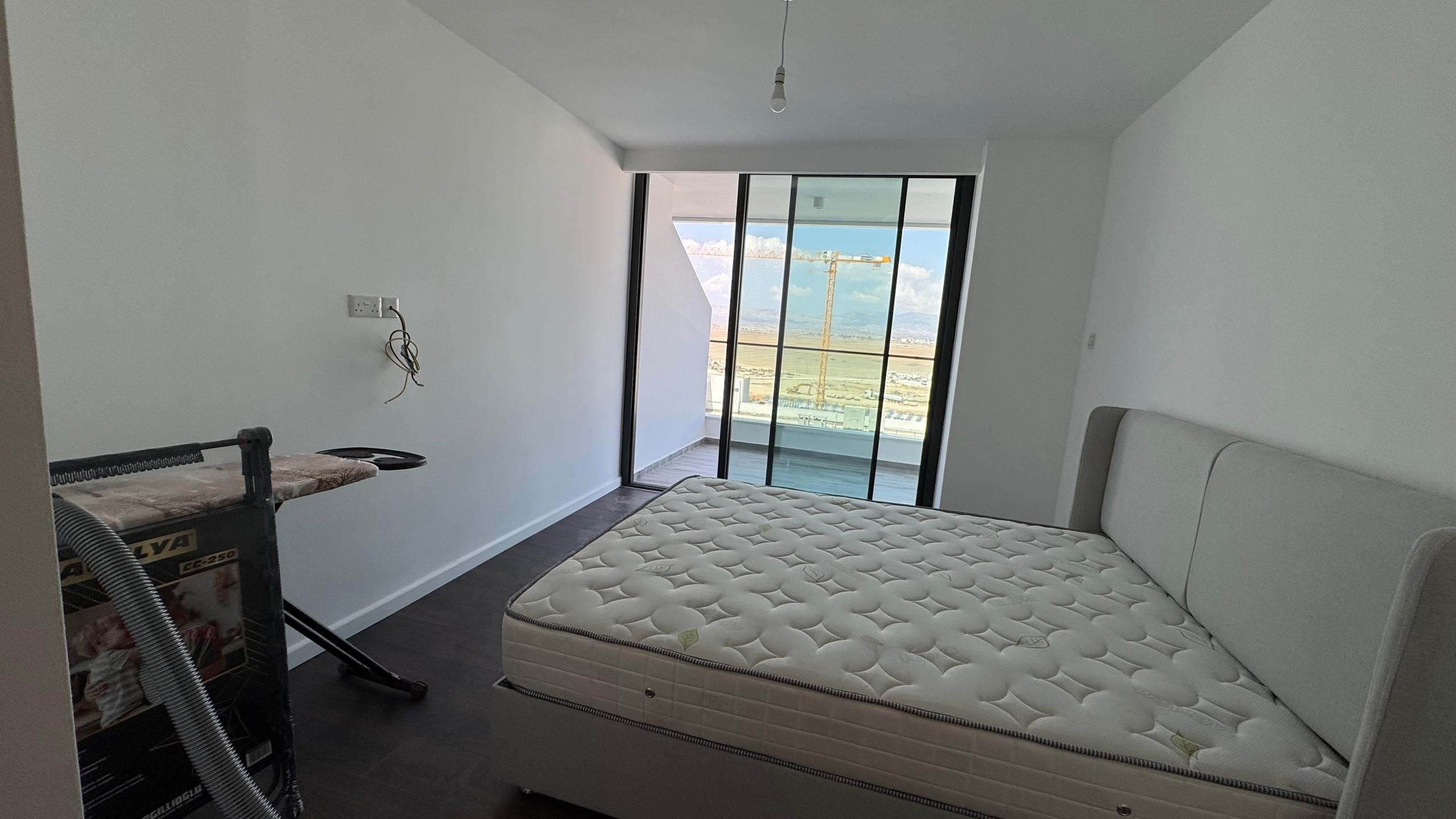 kıbrıs Grand Sapphire Resort & Residence Kıbrıs'ın minimal yatak odasında çıplak bir yatak, ütü masası, beyaz duvarlar, koyu zemin, çıplak ampul ve muhteşem dağ ve gökyüzü manzaralı bir balkona açılan büyük cam kapılar bulunmaktadır. aysan_giti