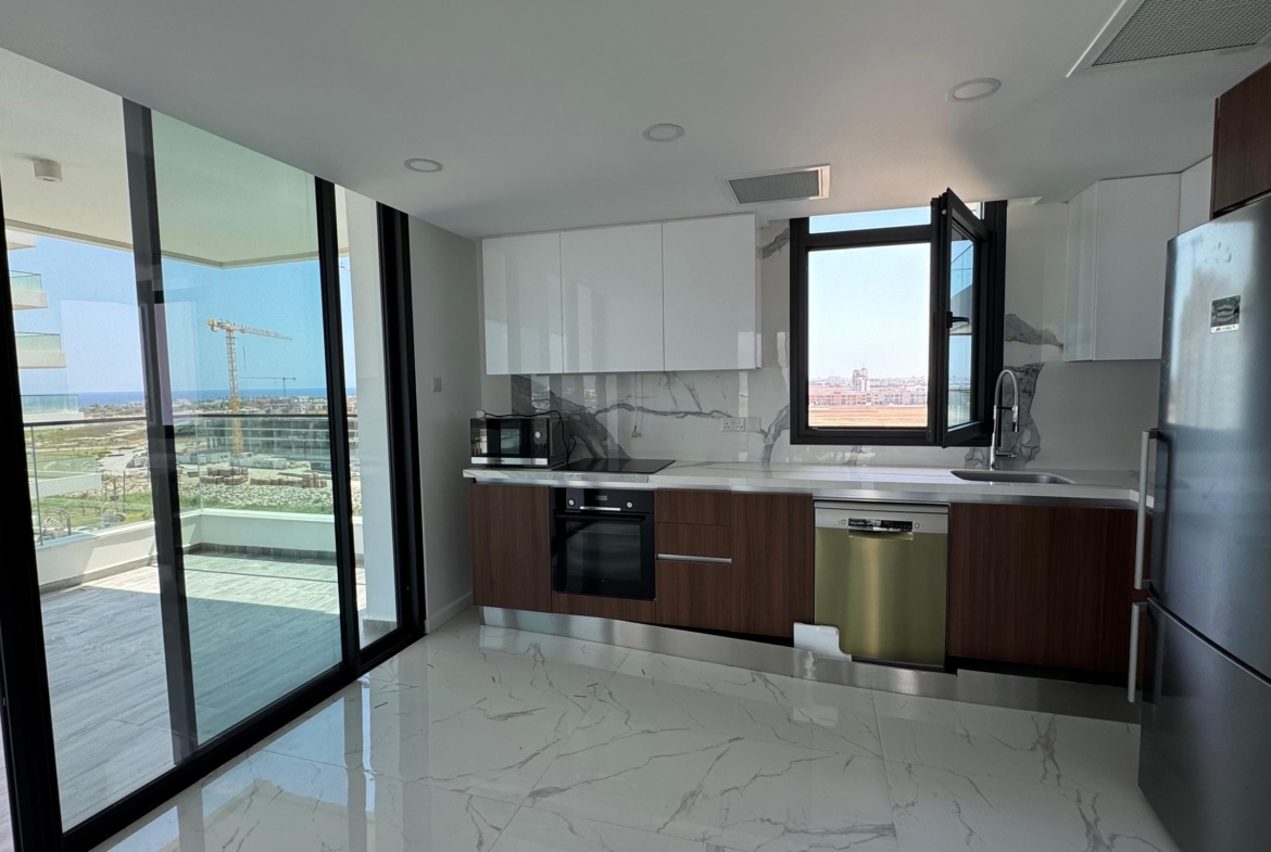 kıbrıs Beyaz ve ahşap dolaplar, paslanmaz çelik aletler, mermer zemin ve açık pencereli modern mutfak. Büyük cam kapılar, Grand Sapphire Resort & Residence'ta Kıbrıs İskele lüks rezidans manzaralı bir balkona açılmaktadır. aysan_giti