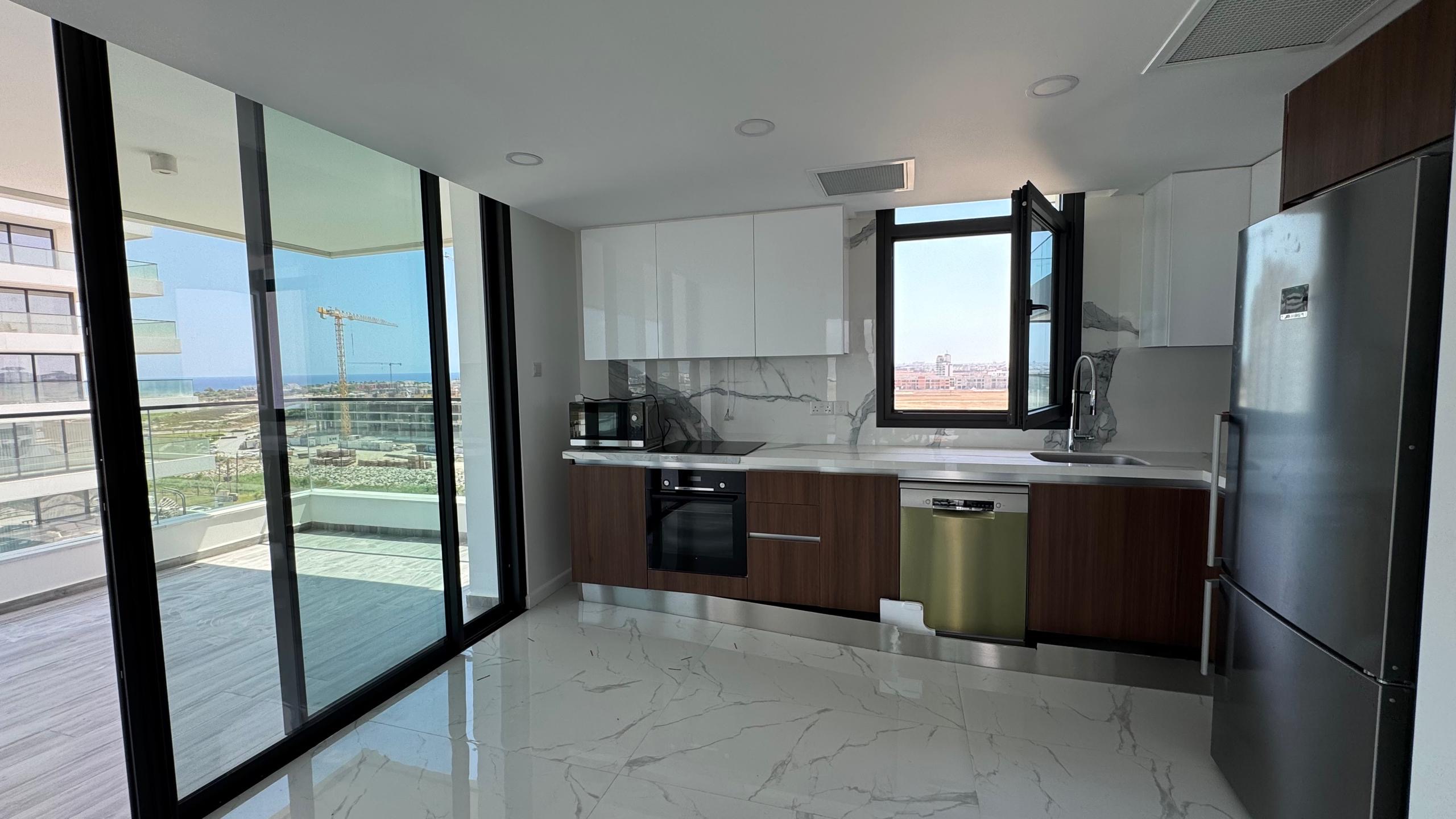 kıbrıs Beyaz ve ahşap dolaplar, paslanmaz çelik aletler, mermer zemin ve açık pencereli modern mutfak. Büyük cam kapılar, Grand Sapphire Resort & Residence'ta Kıbrıs İskele lüks rezidans manzaralı bir balkona açılmaktadır. aysan_giti