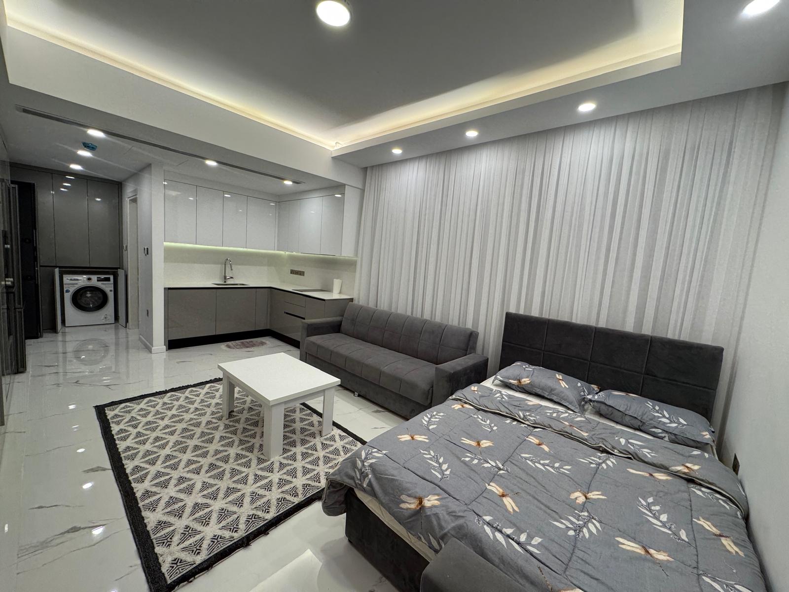 kıbrıs Parlak beyaz zeminli modern stüdyo daire, çiçekli nevresimli gri bir yatak, gri bir kanepe, beyaz bir sehpa, desenli bir halı ve beyaz dolaplar ve ünite içi çamaşır makinesi içeren kompakt bir mutfak alanı. aysan_giti kıbrıs Parlak beyaz zeminli modern stüdyo daire, çiçekli nevresimli gri bir yatak, gri bir kanepe, beyaz bir sehpa, desenli bir halı ve beyaz dolaplar ve ünite içi çamaşır makinesi içeren kompakt bir mutfak alanı. aysan_giti