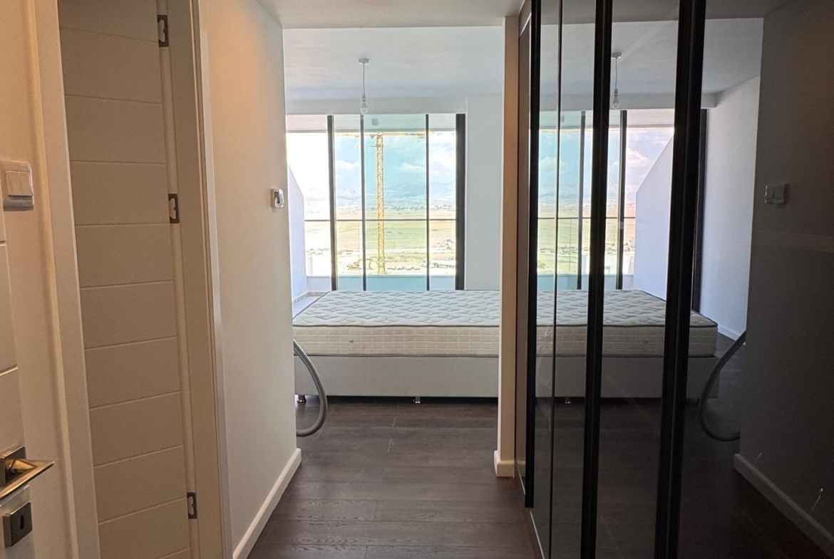 kıbrıs Kıbrıs İskele lüks rezidans Grand Sapphire Resort & Residence'ta koyu ahşap zeminli modern bir koridor, sağda aynalı dolaplar ve ileride deniz ve gökyüzü manzaralı tavandan tabana pencerelere sahip bir yatak odası. aysan_giti