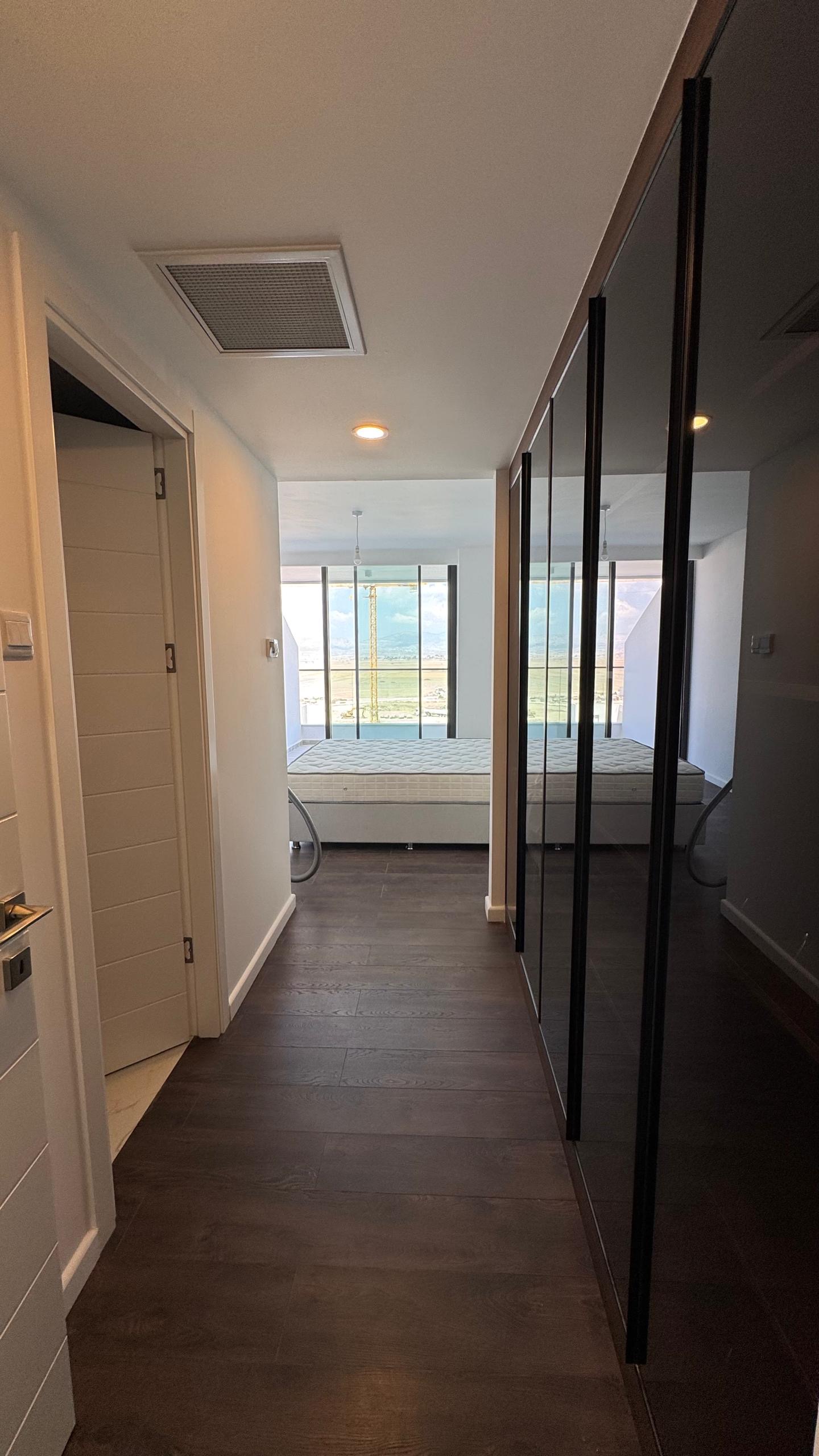 kıbrıs Kıbrıs İskele lüks rezidans Grand Sapphire Resort & Residence'ta koyu ahşap zeminli modern bir koridor, sağda aynalı dolaplar ve ileride deniz ve gökyüzü manzaralı tavandan tabana pencerelere sahip bir yatak odası. aysan_giti