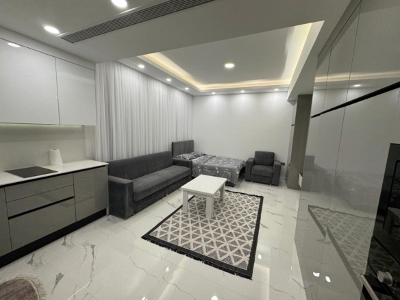 kıbrıs Beyaz duvarları, parlak karo zeminleri, kompakt bir mutfağı, gri bir kanepesi, desenli nevresimli bir yatağı, bir yatar koltuğu, beyaz bir sehpası ve geometrik bir halısı olan modern, minimalist bir stüdyo daire. aysan_giti