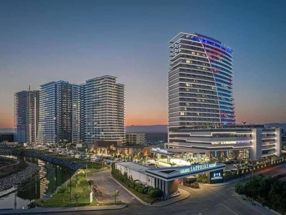kıbrıs Modern yüksek binalar ve alacakaranlıkta ışıklandırılmış Grand Sapphire oteli, küçük bir nehre, sokak lambalarına ve arabalara bakmaktadır. Kuzey Kıbrıs gayrimenkul veya İskele Long Beach kiralık daire arayanlar için mükemmeldir. Gökyüzü gün batımından geceye sorunsuz bir şekilde değişir. aysan_giti