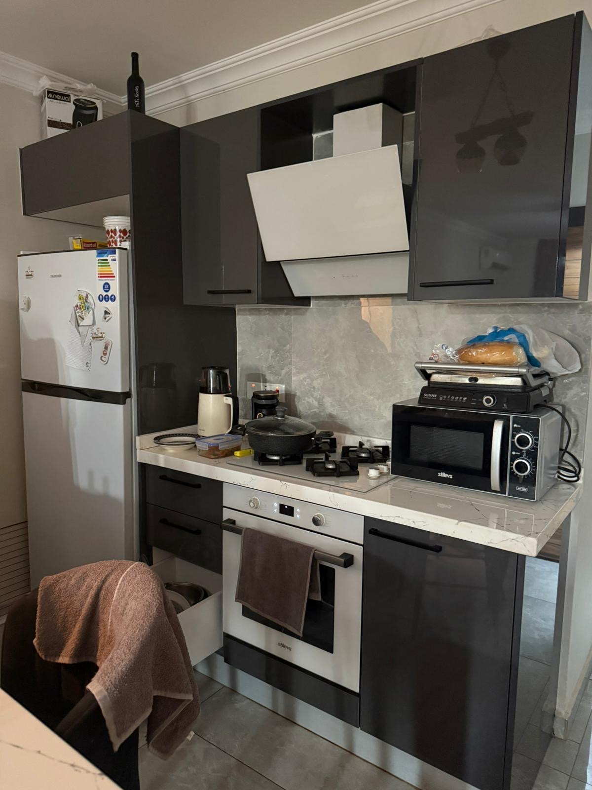 kıbrıs Koyu renk dolapları, beyaz tezgahı ve paslanmaz çelik aletleri olan modern bir mutfak. Tezgâhın üzerinde mıknatıslı bir buzdolabı, fırın, mikrodalga fırın, ekmek kızartma makinesi, su ısıtıcısı ve yiyecek malzemeleri var. Fırından bir havlu sarkıyor. aysan_giti