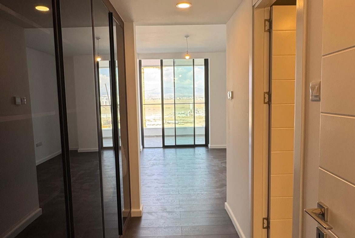 kıbrıs Koyu ahşap zeminli bir koridor, solda aynalı dolap kapıları, sağda beyaz bir kapı ve sonunda doğal ışık alan büyük cam sürgülü kapılar - bir Aysan Giti emlak Kuzey Kıbrıs kiralık daire için mükemmel. aysan_giti