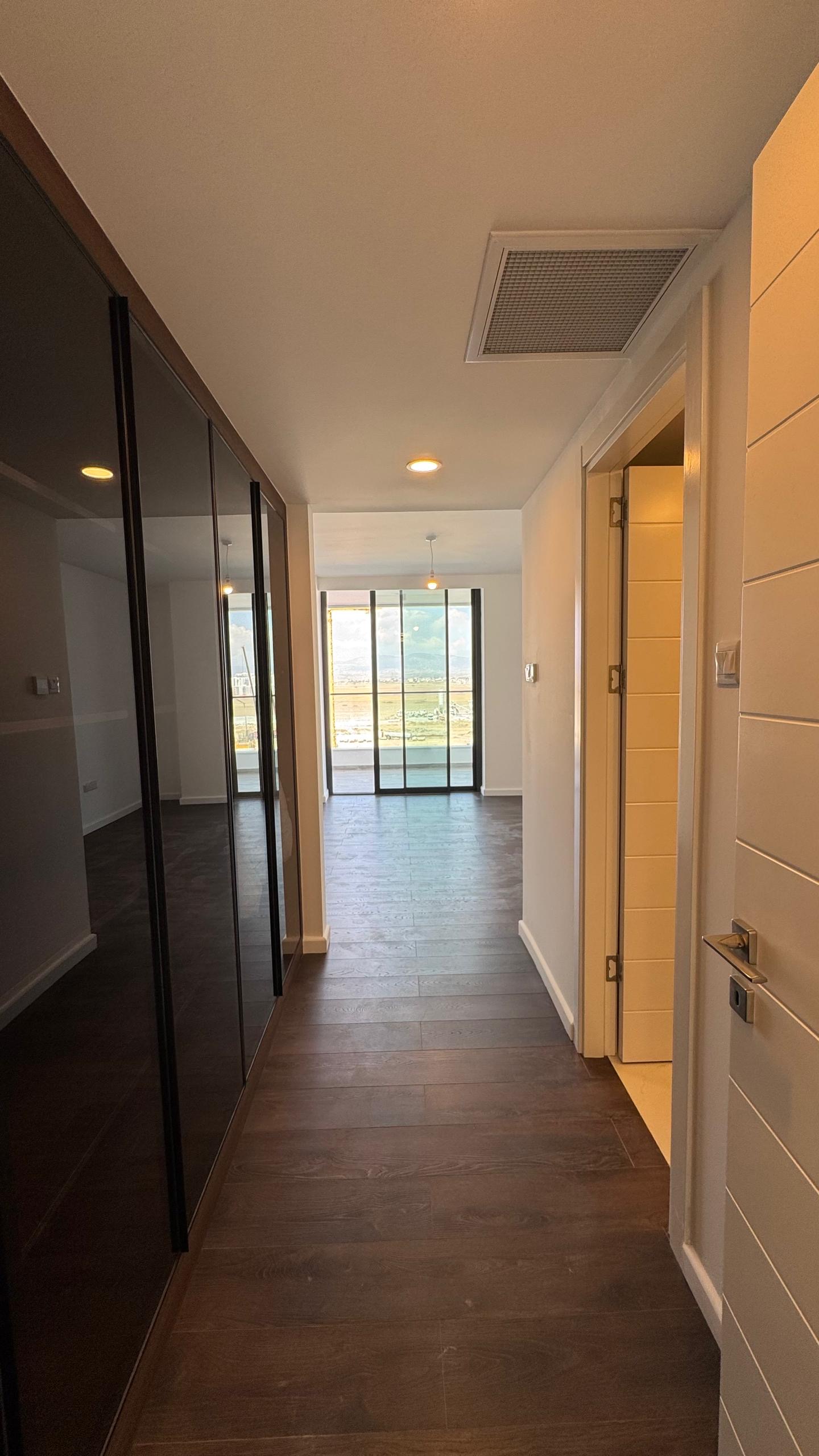 kıbrıs Koyu ahşap zeminli bir koridor, solda aynalı dolap kapıları, sağda beyaz bir kapı ve sonunda doğal ışık alan büyük cam sürgülü kapılar - bir Aysan Giti emlak Kuzey Kıbrıs kiralık daire için mükemmel. aysan_giti