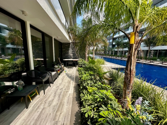 kıbrıs Siyah sandalyeler ve bir masanın bulunduğu modern bir apartman verandası, palmiye ağaçları ve gür yeşilliklerle kaplı, parlak bir gökyüzünün altında çağdaş binalarla çevrili uzun, dar bir yüzme havuzuna bakmaktadır. aysan_giti