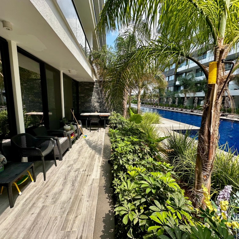 kıbrıs Siyah sandalyeler ve bir masanın bulunduğu modern bir apartman verandası, palmiye ağaçları ve gür yeşilliklerle kaplı, parlak bir gökyüzünün altında çağdaş binalarla çevrili uzun, dar bir yüzme havuzuna bakmaktadır. aysan_giti