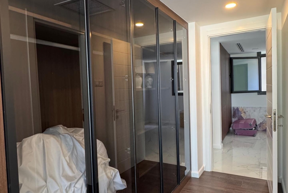 kıbrıs Ahşap zeminli bir koridor ve solda, içinde beyaz yatak takımlarını gösteren cam kapılı bir gardırop - 7 yıldızlı otel konseptinde yaşam Kıbrıs'ı yansıtıyor. Sonunda, açık bir kapı, tavan ışıklarıyla aydınlatılan kutularla döşenmiş bir odayı ortaya çıkarır. aysan_giti