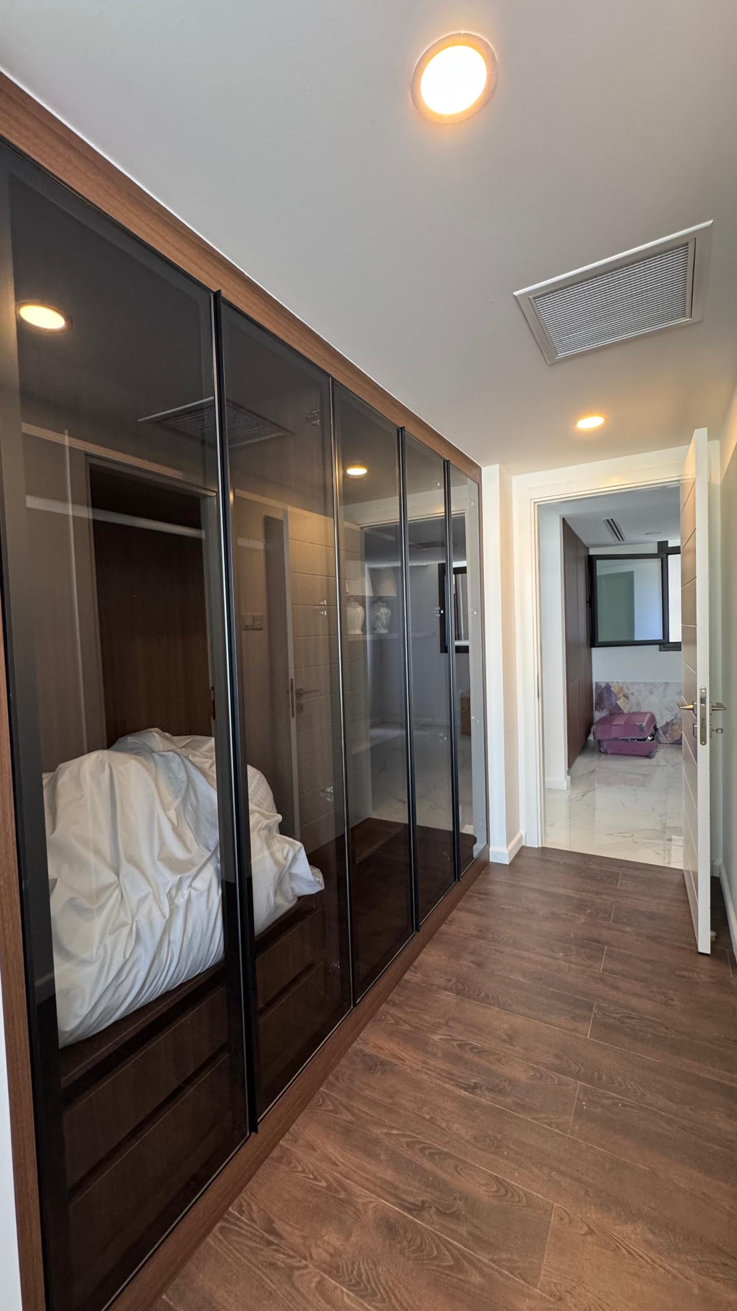 kıbrıs Ahşap zeminli bir koridor ve solda, içinde beyaz yatak takımlarını gösteren cam kapılı bir gardırop - 7 yıldızlı otel konseptinde yaşam Kıbrıs'ı yansıtıyor. Sonunda, açık bir kapı, tavan ışıklarıyla aydınlatılan kutularla döşenmiş bir odayı ortaya çıkarır. aysan_giti