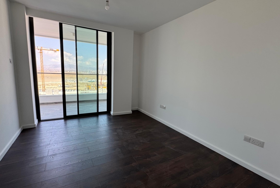 kıbrıs Koyu ahşap zeminli, beyaz duvarlı ve büyük bir cam sürgülü kapısı olan aydınlık, boş bir oda, inşaat alanı, dağlar ve yakındaki lüks Grand Sapphire Resort & Residence manzaralı bir balkona açılıyor. aysan_giti
