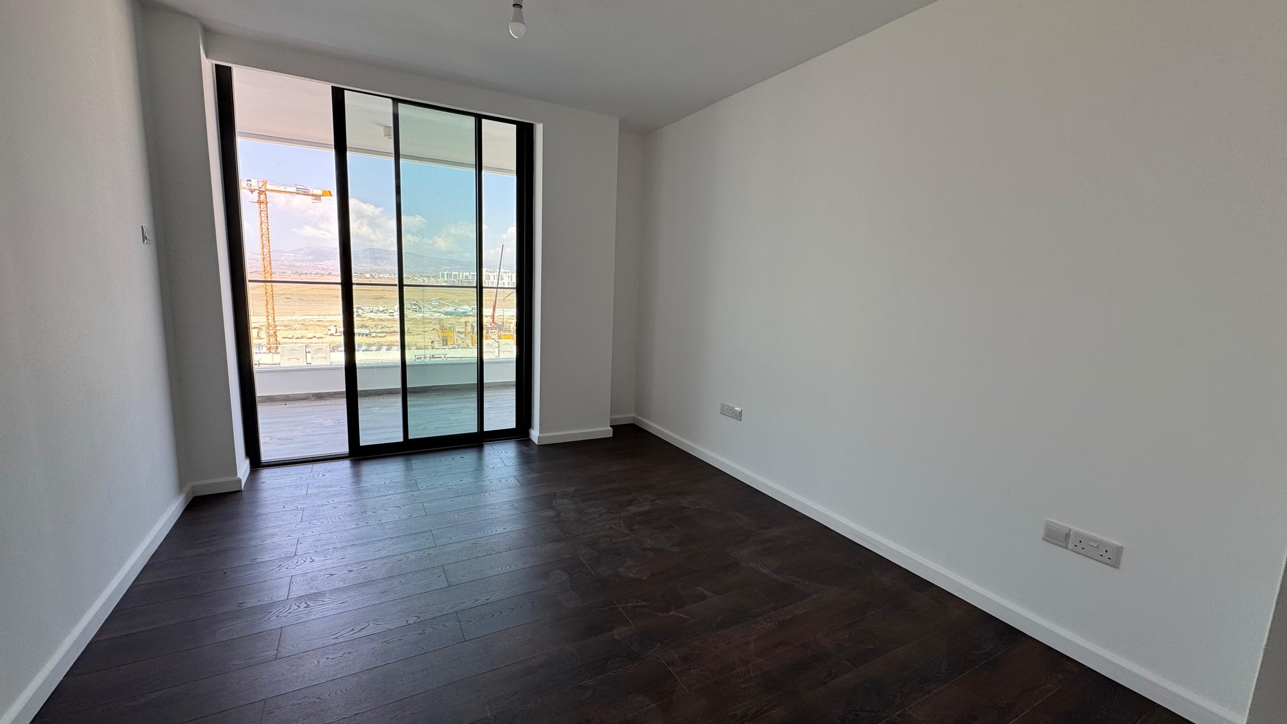 kıbrıs Koyu ahşap zeminli, beyaz duvarlı ve büyük bir cam sürgülü kapısı olan aydınlık, boş bir oda, inşaat alanı, dağlar ve yakındaki lüks Grand Sapphire Resort & Residence manzaralı bir balkona açılıyor. aysan_giti