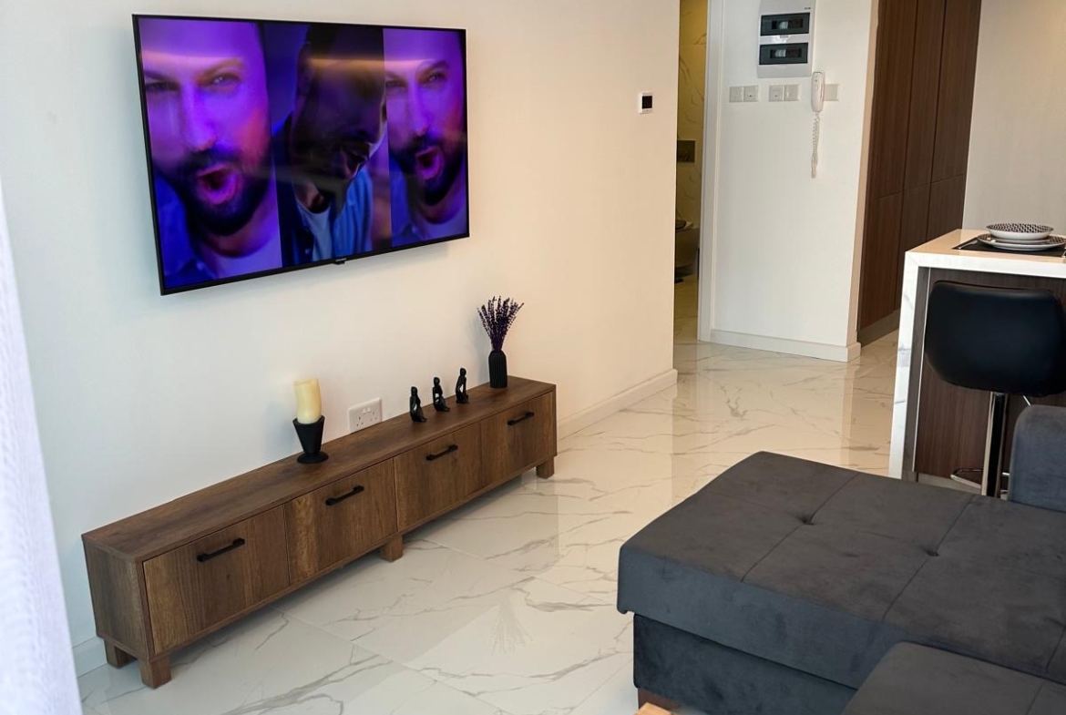 kıbrıs Duvara monte edilmiş bir erkek yüzü gösteren TV, altında ahşap bir TV konsolu ve şık mobilyalarla modern bir oturma odası - Aysan Giti emlak tarafından sunulan Kuzey Kıbrıs'taki İskele Long Beach satılık dairesinin mükemmel bir örneği. aysan_giti