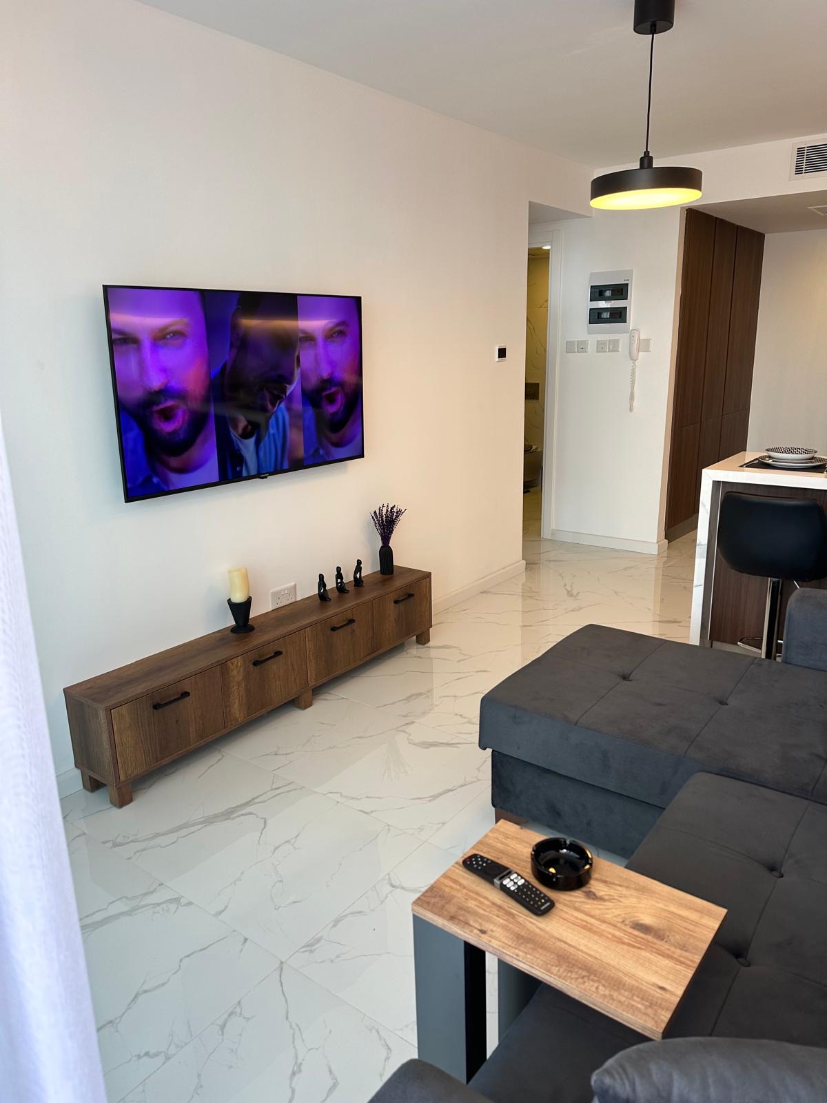 kıbrıs Duvara monte edilmiş bir erkek yüzü gösteren TV, altında ahşap bir TV konsolu ve şık mobilyalarla modern bir oturma odası - Aysan Giti emlak tarafından sunulan Kuzey Kıbrıs'taki İskele Long Beach satılık dairesinin mükemmel bir örneği. aysan_giti